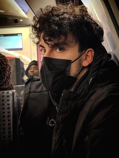 La photographie représente une personne assise dans un train, portant un masque chirurgical noir et une veste noire. La personne a les cheveux bouclés et regarde derrière la caméra. À l'arrière-plan, on aperçoit d'autres passagers et un écran d'information.