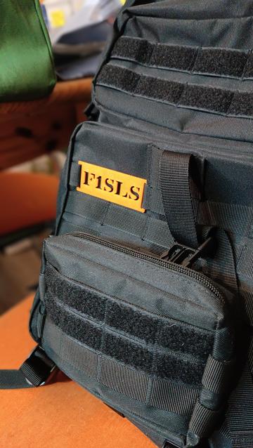 A wide view of the tag installed on a MOLLE strap on my 50L black tactical backpack.

The tag is "F1SLS" written in military style stencil font and appears in black thanks to the negative space.
