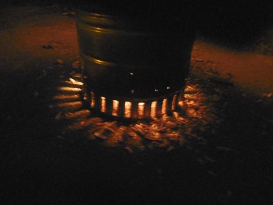 Ein Blechtopf staht auf glattem Boden. Durch eckige Öffnungen scheint das Licht eines Feuers und wirft Schatten in die dunkle Umgebung. Es handelt sich um den Fuß eines Holzgaskochers.