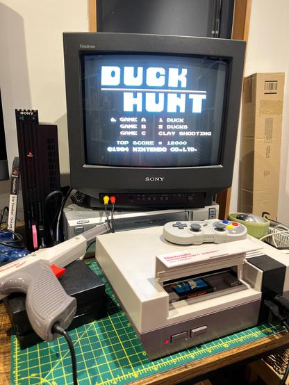 A photo of a Nintendo Entertainment System with the Duck Hunt cartridge inserted. The game’s title screen is displayed on a Sony Trinitron 14” 4:3 CRT TV. a Nintendo Zapper light gun is resting on a small pile of NES cartridges to the right of the NES console. A PS2 can be seen to the left of the TV, which is sitting on top of a Sanyo VHS player.