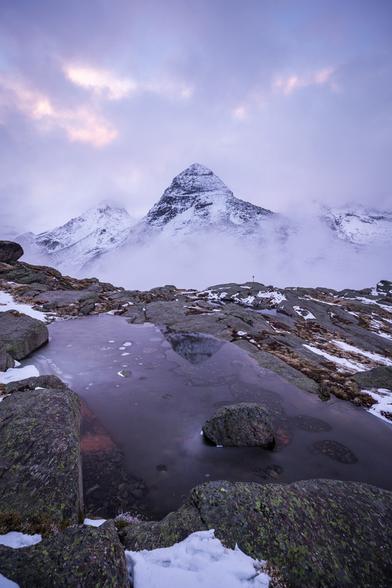 Photo d'un paysage de montagne.
Le premier plan est occupé par une grosse flaque d'eau entre des dalles rocheuses. La surface de l'eau est gelée, donnant un aspect dépoli. Quelques taches de neige apparaissent entre les rochers.
En arrière-plan une montagne à la forme pyramidale et saupoudrée de neige émerge du brouillard.
Le ciel est nuageux, mais au-dessus de la montagne, quelques taches plus claires et colorées donnent un aspect surnaturel.
-
Photo of a mountain landscape.
The foreground is occupied by a large puddle between rocky slabs. The surface of the water is frozen, giving it a frosted appearance. A few patches of snow appear between the rocks.
In the background, a snow-dusted, pyramid-shaped mountain emerges from the fog.
The sky is cloudy, but above the mountain, a few brighter, more colorful patches give an unearthly appearance.