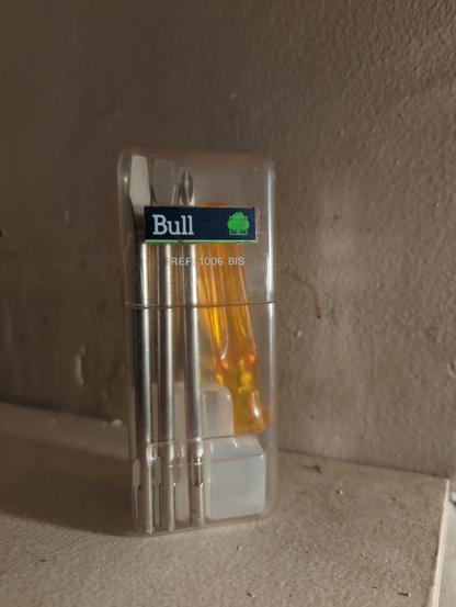 Une boîte en plastique transparent arbore un autocollant avec le logo bleu foncé et vert de Bull. Elle contient des lames de tournevis plats et cruciformes entre 3 et 6 mm et un manche de tournevis en plastique orange transparent.