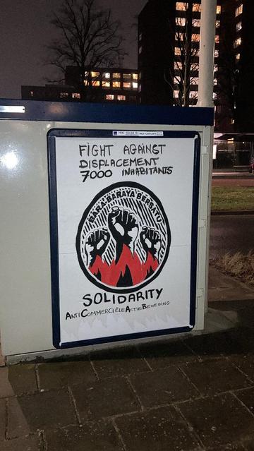 An electricity box at a crossroads on the street at nightlight
with a billboard on it.
White with a black circle with 3 fists in it up in black white with red black flames and the text Bara-Baraya Bersatu.
Above and below text as in caption