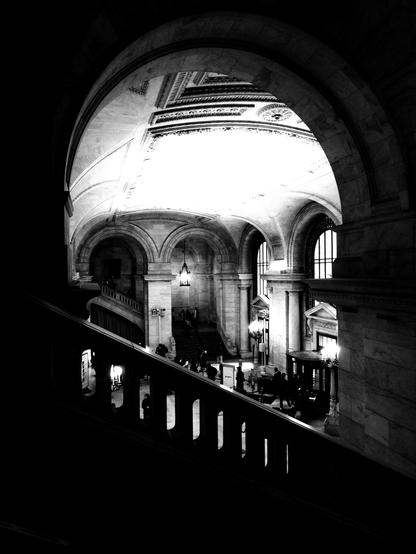 Black and white photograph of the hall of the New York Public Library, taken from one of the side a staircases. The hall is visible from above, through one of the arches.
