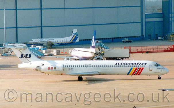 Side view of a white, twin engined jet airliner with the engines mounted on the side of the rear fuselage, taxiing from left to right.
There are a number of coloured near-vertical bars - blue, yellow, red, and black -  on the lower forward fuselage, starting under the passenger cabin windows and looping under the belly, as well as squares on the engine pods - red with a white cross, red with a black cross inside a white cross, and blue with a yellow cross, each representing the countries Sweden, Norway, and Denmark.
There are black "Scandinavian" titles on the upper forward fuselage and a black "SAS" logo on the tail.
Grey concrete fuselage fills the foreground, with another white, twin engined jet airliner with a blue belly and tail in the immediate background, facing more or less away from the camera.
Beyond that, a white, twin propellor-engined airliner with a blue belly and a yellow stripe running along the body is parked facing to the left in front of a large grey hangar, the hangars doors dwarfing this latter plane.