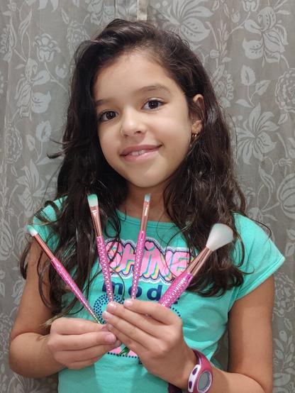 A young girl named Sarah smiles at the camera, holding four pink makeup brushes in her hands. She has long, dark, wavy hair and is wearing a light green t-shirt with the words "SHINE mode" written on it. The brushes have sparkly pink handles. The background is a curtain with a gray and white floral pattern.