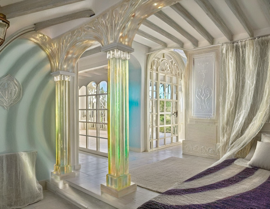 A dreamy bedroom with glowing green and gold tinted glass columns, gossamer white curtains, a bed with a purple and white chenille spread, and a window and door overlooking a sunny patio.