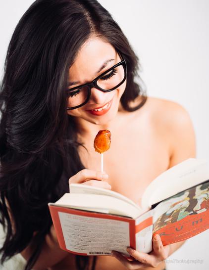 Image of a young lady with long hair and pretend glasses holding a Rumi book and a lollipop. She is half smiling and half laughing. Her eyes are down at the book.