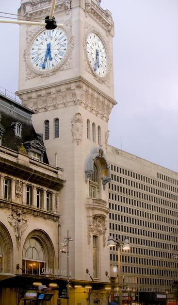 La photographie représente la tour de l'horloge de la gare de Lyon à Paris. La tour présente une architecture ornée de cadrans d'horloge sur plusieurs côtés. Le bâtiment de la gare est visible dans la partie inférieure de l'image, y compris une partie de sa façade détaillée et de ses éléments décoratifs. Un autre bâtiment est visible sur la droite de l'image, avec une architecture neutre, un bloc de béton avec plein de fenêtre. La différence d'architecture crée un contraste dans l'image.