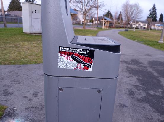 A rectangular sticker that says "Trans People's Joy Enriches Everyone. A Society That Represses It Stiffles Us All" with art of a boot about to crush a flower. The sticker is on a water fountain next to a trail in a city park and the picture was taken close to sunset.