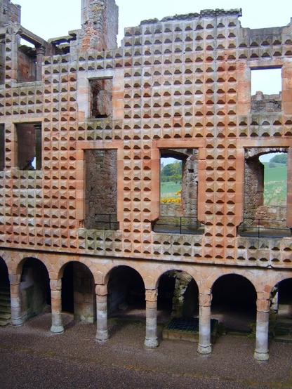 The interior facade of Crichton Castle, Scotland, featuring a textured wall with a distinctive diamond-patterned facade, upper level openings, and a lower level arcade with rounded arches and columns.