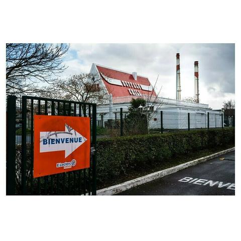 Entrée d’un parking avec une grande flèche orange portant l’inscription « Bienvenue – E.Leclerc Drive ». En arrière-plan, un bâtiment blanc au toit rouge et deux cheminées industrielles se détachent sous un ciel nuageux, créant un contraste entre l’aspect commercial et l’environnement industriel.