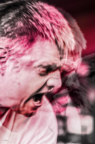 Colour photo in red/rose spectrum, doible exposure of the head of a person who is yelling at another person in furious anger (the other person is not visible in the picture), eyes narrowed into slits, mouth as if drooling, face twisted into angry lines.