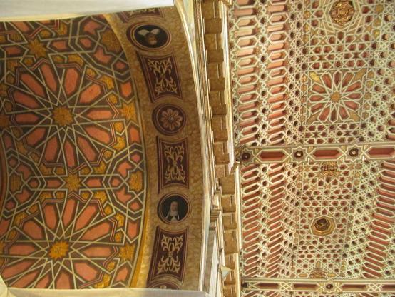 Photo of a richly decorated ceiling. On the left are 12-pointed rosettes. In the middle is an arch with floral and vegetal ornaments and two portraits. On the right are 6-, 8- and 12-pointed stars. Red, white and gold colours dominate.