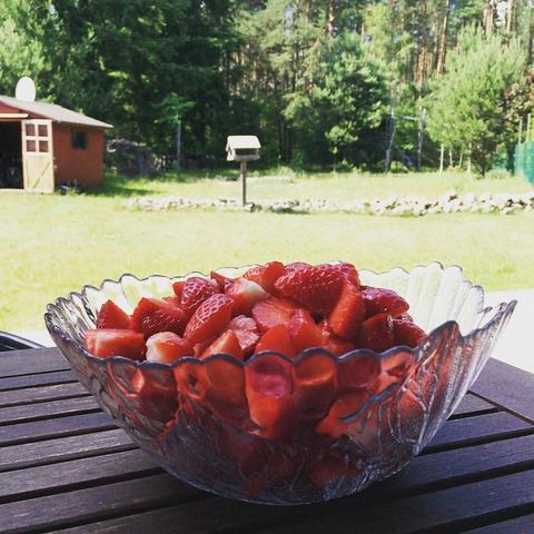 A glass bowl filled with sliced strawberries sits on a wooden table outdoors. In the background, there is a grassy lawn, a small shed, and trees.
