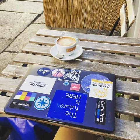 A wooden outdoor table with a laptop covered in various colorful stickers and a cappuccino in a white cup on a saucer. The space is partially enclosed by a bamboo fence, and there's a hint of a chair visible.