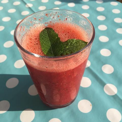 A glass filled with a frothy red smoothie, garnished with fresh mint leaves, sits on a blue tablecloth with white polka dots.