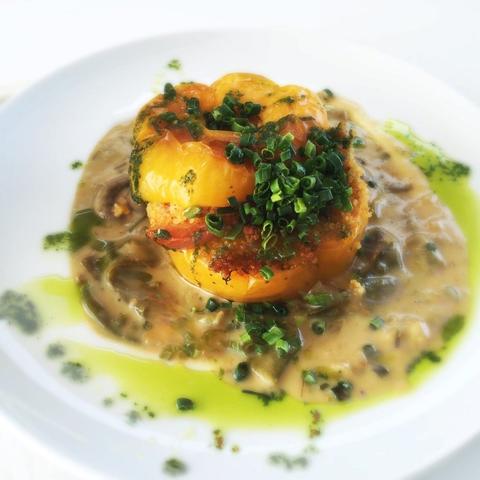 A vibrant yellow bell pepper stuffed with a textured filling, garnished with chopped green herbs, sits atop a creamy sauce drizzled with green oil on a white plate.