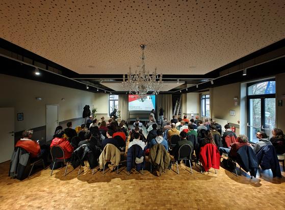 A conference room filled with people sitting on chairs, facing a presentation screen. The room features a crystal chandelier, parquet flooring, and a perforated ceiling. The audience is wearing winter coats in various colors including red, blue, and beige.