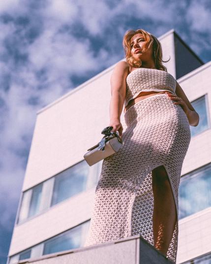 A confident woman in a beige crochet two-piece outfit stands elegantly against a modern white building with a blue sky backdrop. She holds a stylish handbag while sunlight highlights her features, creating a powerful and sophisticated urban fashion aesthetic.