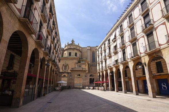 A empty public square in a European city, almost universally light, warm-coloured stone. It's a long but not very wide space with square paving stones laid diagonally across the open area. Buildings either side of the long end are five storeys high with the upper three storeys supported by an arcade with stone blocks curved with Romanesque arch openings. In the shade under the arcade on the left there is seating for a café or restaurant, while on the right we can shop fronts with shutters down at the moment. At the far end of the more small tables and chairs with parasols have been set up either side of a low wall with large square opening in it to allow egress from this space. Beyond the square another building in the same yellow-white stone as we see here stretches across the gap; a tall window and octagonal tower structure to the left suggest it is a medieval church.