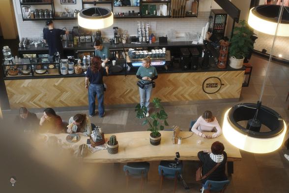 The image shows the interior of a coffee shop from above. There is a long wooden table with people sitting and working or conversing. The counter is staffed by baristas, and you can see various coffee-making equipment on it. The interior is well-lit with large, round hanging lights, and there are some plants and decor around.