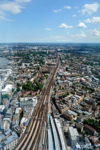 An aerial photograph shows a section of London on a sunny day, with numerous buildings alongside the railway tracks leading into London Bridge Station. Several trains are visible on the tracks. The sky is blue with scattered white clouds.