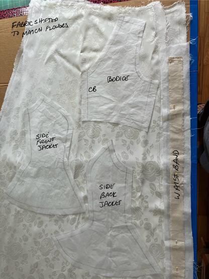 Jaquard fabric in ivory laid on a cardboard butting board with white tissue paper patterns distriubuted on top. A bodice, afew jacket patterns and a waistband.