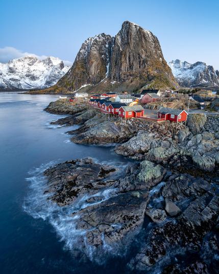 The image features a scenic coastal landscape with red wooden houses, typical of fishing villages, situated on rocky shores. In the background, there are large, dramatic snow-covered mountains. The water is calm, and the sky is clear, suggesting a serene and picturesque environment, likely in a northern region such as Norway.