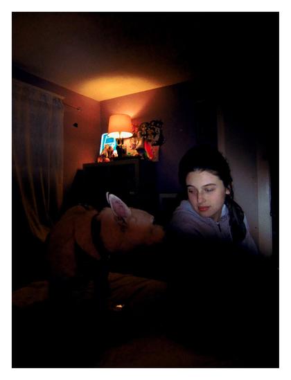 an amateur photograph of a girl seated next to a dog in bed. she looks at it with an admiring smile on her face. The room is dark, the only lights a small lamp in the corner and a suppressed flash which casts over the girl's face, revealing her expression. the dog is still shrouded in dark.