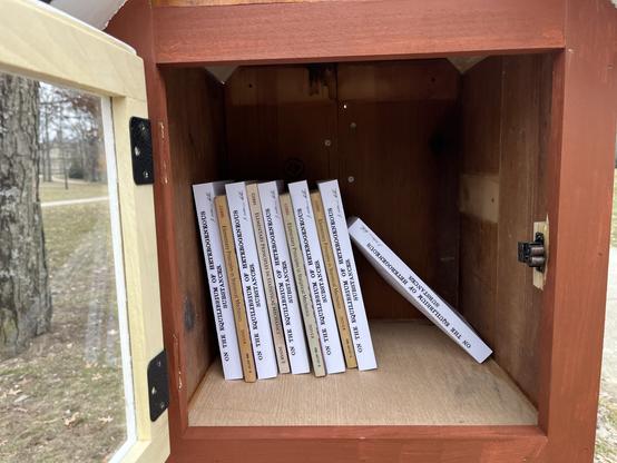 A close photo of a painted wooden box with an open, clear door, with tended grass and trees in the background. In the box are a mixture of ~8.5" tall books, spines facing the viewer. They are either white, with the title "ON THE EQUILIBRIUM OF HETEROGENEOUS SUBSTANCES" with a script signature (J. Willard Gibbs) or tan with "GIBBS      Elementary Principles in Statistical Mechanics". There are 10 book, which ~1/2 fills the box
