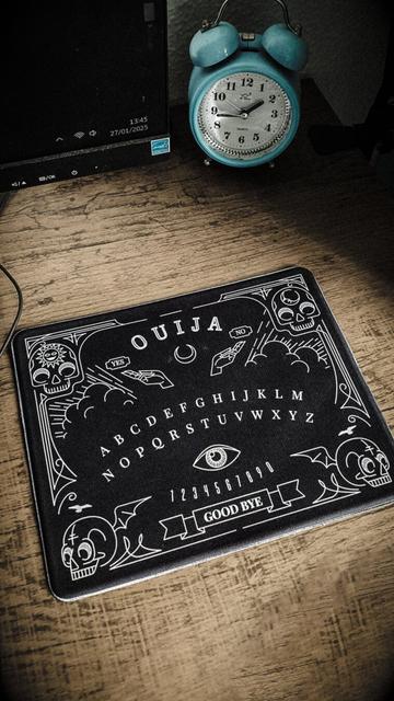A imagem mostra um ambiente de trabalho ou estudo com um toque misterioso. Sobre uma mesa de madeira, há um tapete de mouse estilizado como um tabuleiro de Ouija, com ilustrações de mãos, crânios, luas, estrelas e elementos místicos. Ele apresenta as palavras "YES" e "NO" no topo, o alfabeto no centro e os números de 0 a 9, além da inscrição "GOOD BYE" na parte inferior.

Ao fundo, há um despertador retrô azul, marcando 13 e 45 aproximadamente. À esquerda, parte de um monitor de computador está visível, exibindo a hora 13h45 e a data 27/01/2025. A iluminação é suave, criando sombras e um tom levemente sombrio na cena. O estilo da foto remete a uma estética vintage e esotérica.