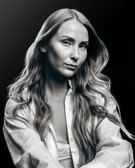 A black and white fine art portrait of a woman with long, wavy hair. She gazes confidently at the camera, wearing a white button-down shirt over a lace top. The soft lighting enhances the contours of her face and the delicate details of her clothing, creating a timeless and elegant composition.
