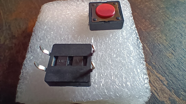 A couple of replacement switches for old DOD/Digitech guitar effect pedals that I got from 'Bay or Reverb a while ago, sitting on some white plastic foam.
One is button-side-up, and one is leg-side-up, showing four legs to wire into the pedal.
They are a bit of a pain to replace, and they go bad every few years.
Grr.