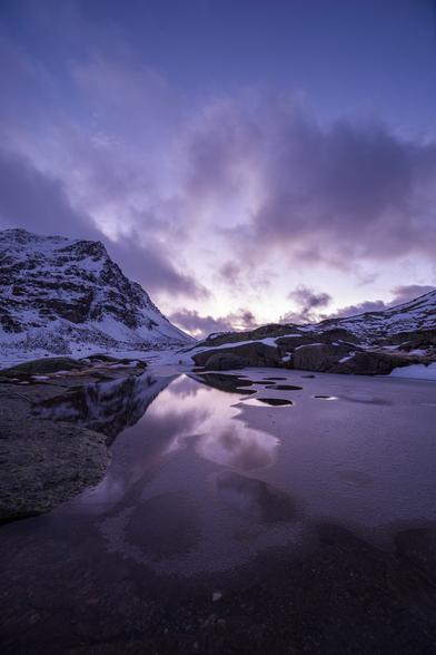 Photo d'un paysage de montagne.
Au premier plan se trouve un lac partiellement gelé, certaines zones d'eaux libres forment des ronds dans la glace.
Le paysage autour du lac est saupoudré de neige.
Le ciel est clair mais à moitié ennuagé, la lumière vive et jaunâtre à l'horizon indique que le soleil vient de se coucher. Les couleurs violacées du ciel se reflètent dans le lac.
-
Photo of a mountain landscape.
In the foreground is a partially frozen lake, with some areas of open water forming circles in the ice.
The landscape around the lake is dusted with snow.
The sky is clear but half clouded, and the bright, yellowish light on the horizon indicates that the sun has just set. The purplish colors of the sky are reflected in the lake.