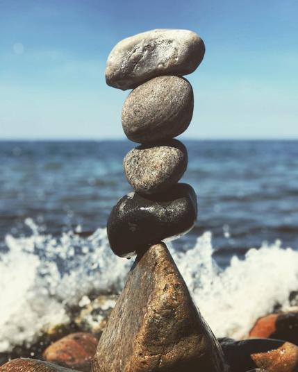 Four beach rocks balancing on top of each other over a larger stone. In the background crashing waves are incoming over the spread out rocks in the background.