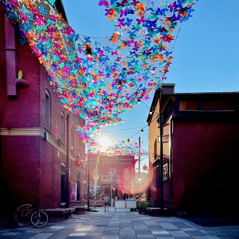 The image depicts a vibrant alleyway adorned with colorful butterfly decorations hanging from a net above. The scene is illuminated by sunlight, creating a warm and inviting atmosphere. On either side, there are brick buildings, and the ground is paved with large tiles. In the background, a few people can be seen enjoying the space, adding to the lively ambiance.