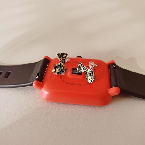 a pair of small stud earrings with an old tiny PCB with black and copper colors. they are placed on an orange smartwatch with black silicone band