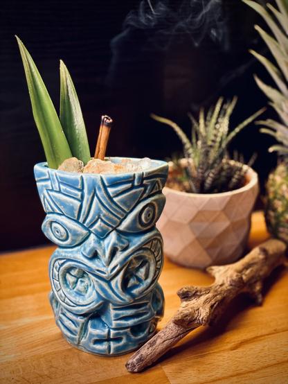 cocktail with pineapple frond and burnt cinnamon stick garnish still smoking inside a blue ceramic Bosko tiki mug with driftwood in the foreground and cacti and pineapple in the background