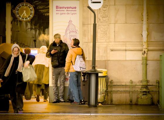 La photographie représente un groupe de personnes devant une gare, à côté de son entrée. Il y a au-dessus d'eux un panneau qui dit « Bienvenue en gare de Lyon ». On aperçoit à travers l'entrée une horloge suspendue. Deux personnes sont debout sous le panneau, à côté de poubelles et d'un panneau avec une cigarette suggérant un espace fumeur l'une d'elle fume avec un casque sur les oreilles, un nuage de fumée s'échappe de sa bouche, l'autre consulte son téléphone portable. Une femme semble quant à elle sortir de la gare, marchant tête baissée vers l'objectif, avec sa valise. La colorimétrie est chaude, avec beaucoup de jaune ponctué par du gris.