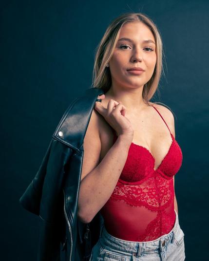 The image shows a person with long hair wearing a red lace top, denim shorts, and holding a black leather jacket over their shoulder. The background is dark.