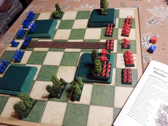 A large green and white chessboard adorned with green square hills, a brown road down the center, and some stands of suitably small trees, with blue and red Risk pieces (cavalry, cannon, and infantry in the Napoleonic style) arrayed at opposite ends of the board.