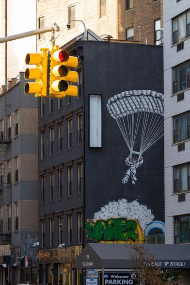View over a street in New York. On the other side, a black house gable with a parachutist sprayed on it in white paint. On the left in the foreground, one of the typical yellow traffic lights