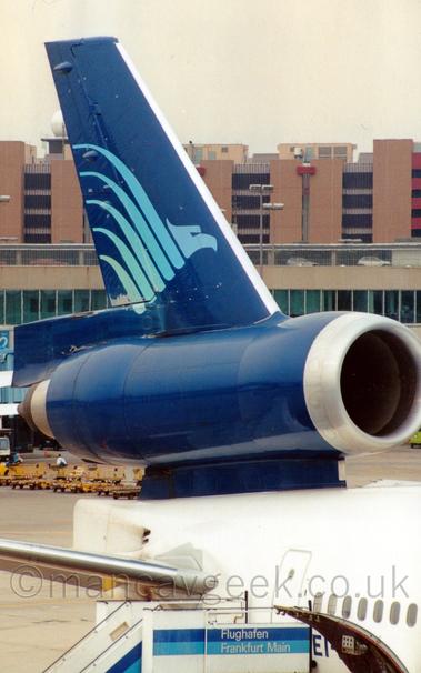 Close-up of the dark blue tail and centre engine of a white, 3 engined jet airliner parked facing to the right.
There is a stylised, light blue flying bird with wings stretched out on the tail.
A set of mobile airstairs is attached to the closed rear door under the tail, with a pair of dark and light blue stripes with white writing "Flughafen Frankfurt Main"  on the side.
A door in the loser fuselage is hinged to open upwards, partially obscuring the planes registration marks, so only "EI-" is visible.
In the background, Brown and grey buildings stretch across the frame, under grey skies.