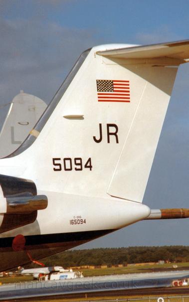 Closeup of the white tail and rear fuselage of a twin engined BizJet with the rear of the engines visible mounted on the sides of the rear fuselage, parked facing to the left.
There is a US flag at the top of the tail, with large letters "JR" in black underneath, and the number "5094" below that and very slightly further forward.
The tailcone has text "C-20G" in very small letters, with the number "165094" in slightly larger letters below that.
A black stripe runs along the body, wrapping around under the tailcone.
The planes wing stretches across the bottom of the frame, with a white, twin engined jet airliner visible over the top, with trees filling the skyline , under blue-grey skies.