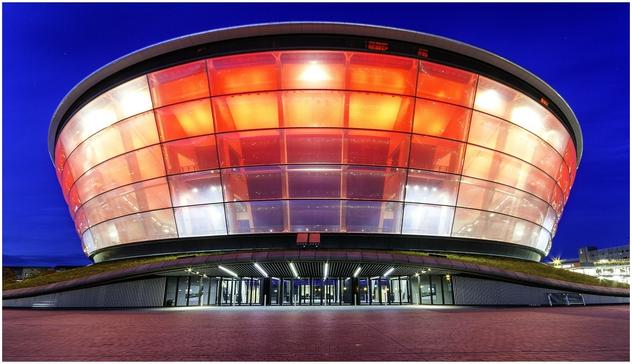 A modern stadium with a circular design, featuring a transparent upper section illuminated in red against a twilight blue sky. The entrance has revolving doors and a sleek, contemporary façade.