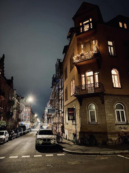 Gerne! Hier ist eine Bildbeschreibung des Fotos, das Sie geteilt haben:
Einleitung:
Das Foto zeigt eine nächtliche Straßenszene in einer Stadt, aufgenommen im Hochformat. Es ist dunkel, aber die Straßenlaternen und die Lichter der Gebäude erhellen die Szene. Das Bild hat eine ruhige und fast melancholische Atmosphäre.
Hauptteil:
Im Vordergrund des Bildes befindet sich eine dunkle, leere Straße, die von zwei Reihen geparkter Autos gesäumt wird. Die Autos sind in verschiedenen Farben und Modellen zu sehen. Die Straße ist sauber und gut beleuchtet, mit einer durchgezogenen Linie in der Mitte. Am Ende der Straße befindet sich eine Kreuzung, an der ein einzelnes Auto mit eingeschalteten Scheinwerfern steht und nach links abbiegt.
Auf beiden Seiten der Straße befinden sich mehrstöckige Gebäude mit vielen Fenstern. Die meisten Fenster sind dunkel, aber einige sind beleuchtet und geben einen Einblick in die Innenräume der Wohnungen. Die Gebäude sind aus verschiedenen Materialien gebaut, darunter Ziegel und Stein, und weisen unterschiedliche architektonische Stile auf. Einige Gebäude haben aufwendige Details wie Erkerfenster und Balkone.
Der Himmel über der Straße ist dunkel und bewölkt. Es sind keine Sterne oder Monde zu sehen. Die Wolken verleihen dem Bild eine dramatische und düstere Note.
Zusammenfassung:
Das Foto ist ein gutes Beispiel für die Nachtfotografie in Städten. Es fängt die Stille und Einsamkeit einer nächtlichen Straße ein, während es gleichzeitig die Schönheit der stä