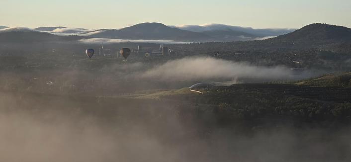 A pair of balloons float above some clearing mist. The armadillo shaped arboretum pavilion is below with the green hill side lit up by the morning sun.