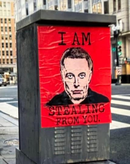 A bold red street art poster featuring a stylized black-and-white portrait of Elon Musk is pasted onto a utility box in an urban setting. The text above his image reads "I AM," while the text below states "STEALING FROM YOU." The striking contrast between the red background and the black typography enhances the provocative message. Surrounding the scene are tall office buildings and a city intersection, indicating a downtown area. The poster appears slightly wrinkled, suggesting it was recently put up.