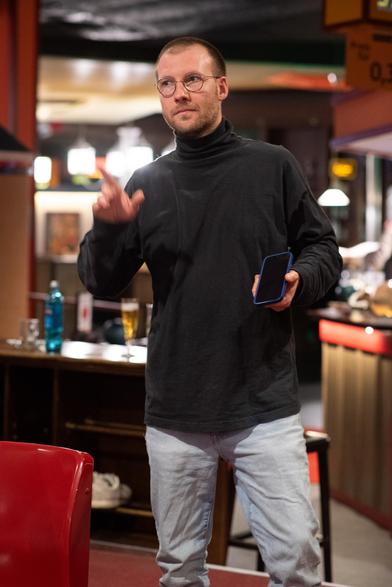 A man dressed in a black turtleneck and light-colored Jeans, stands within a dimly lit Establishment. The room is reminiscent of a retro bar or lounge.